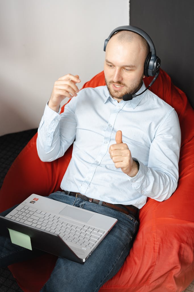 Caucasian man in white shirt using laptop with headset on beanbag chair, engaging in remote work.