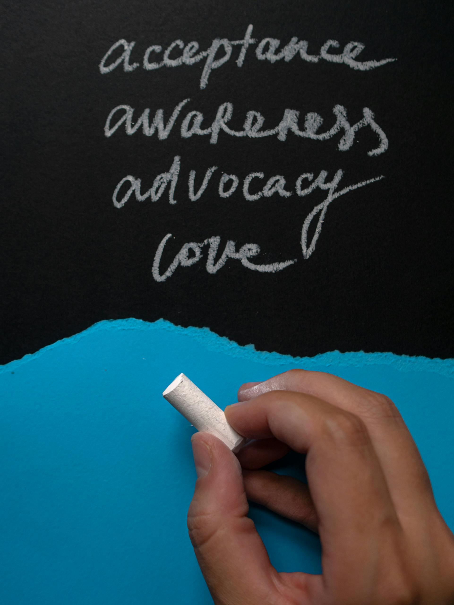 Hand with chalk writing words of acceptance, awareness, advocacy, and love on blackboard.